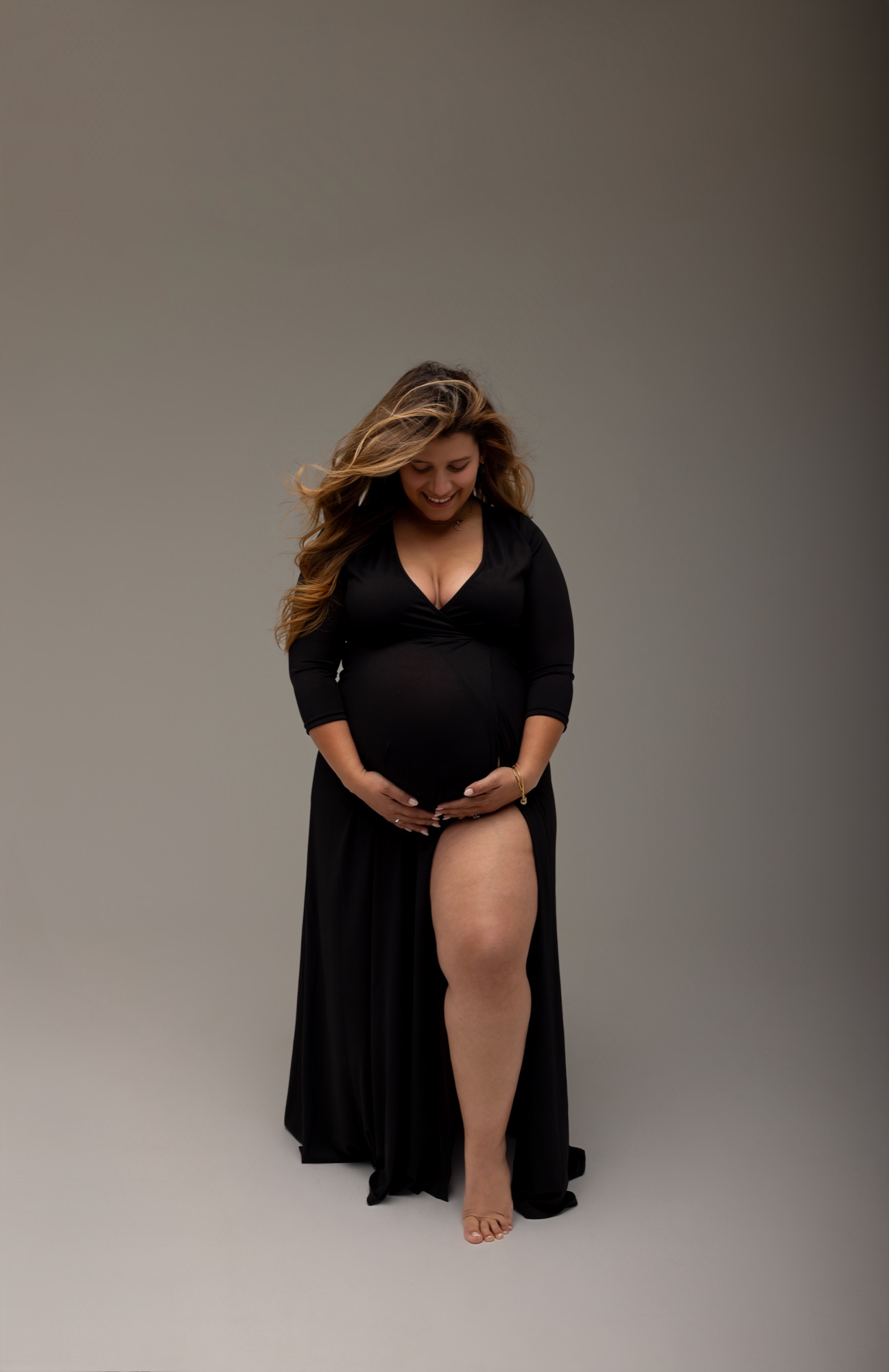 Pregnant woman in a black gown smiling down at her hands on her bump against a grey studio backdrop