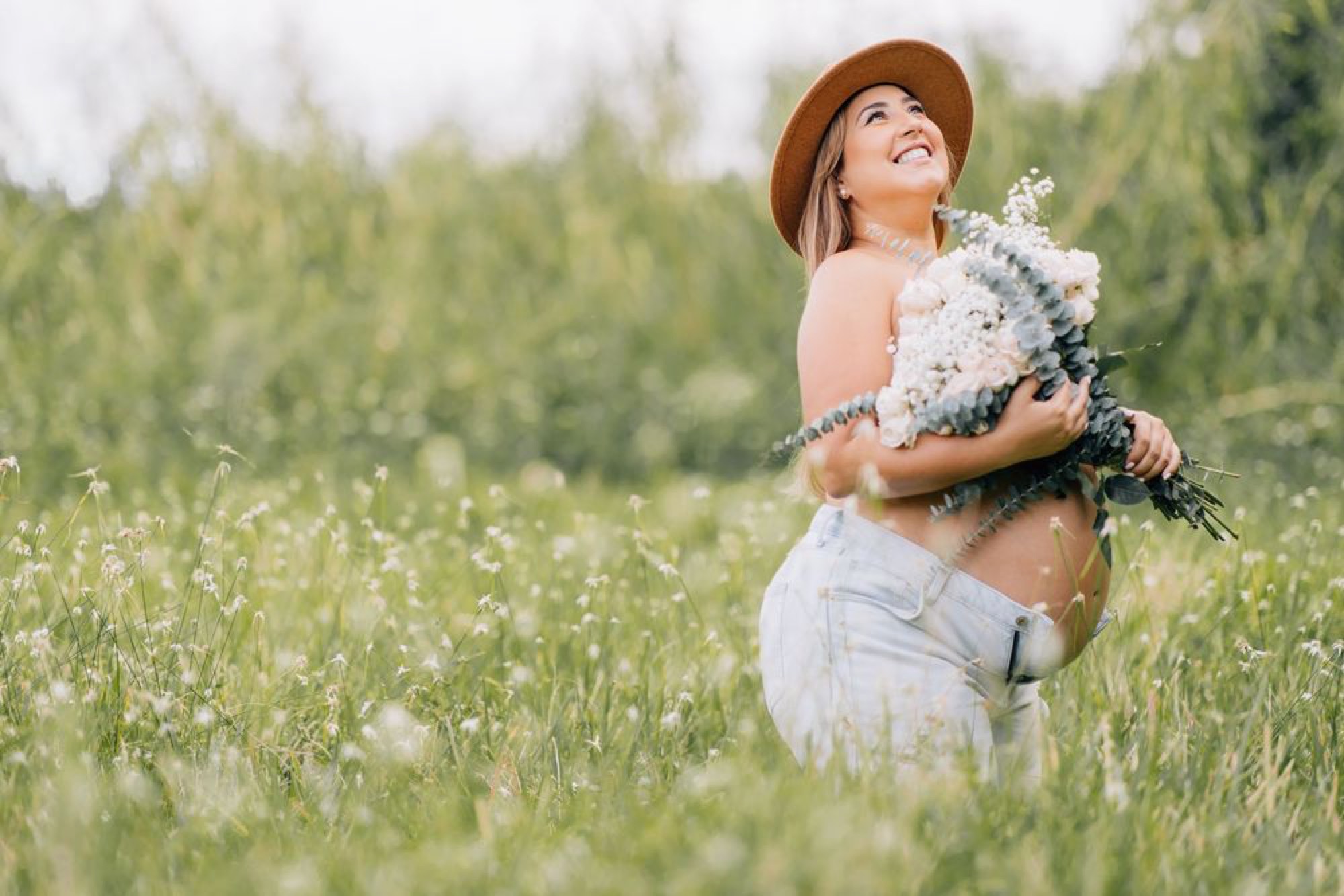 Maternity portrait in a sunlit meadow, bouquet in hand and face turned to the sky