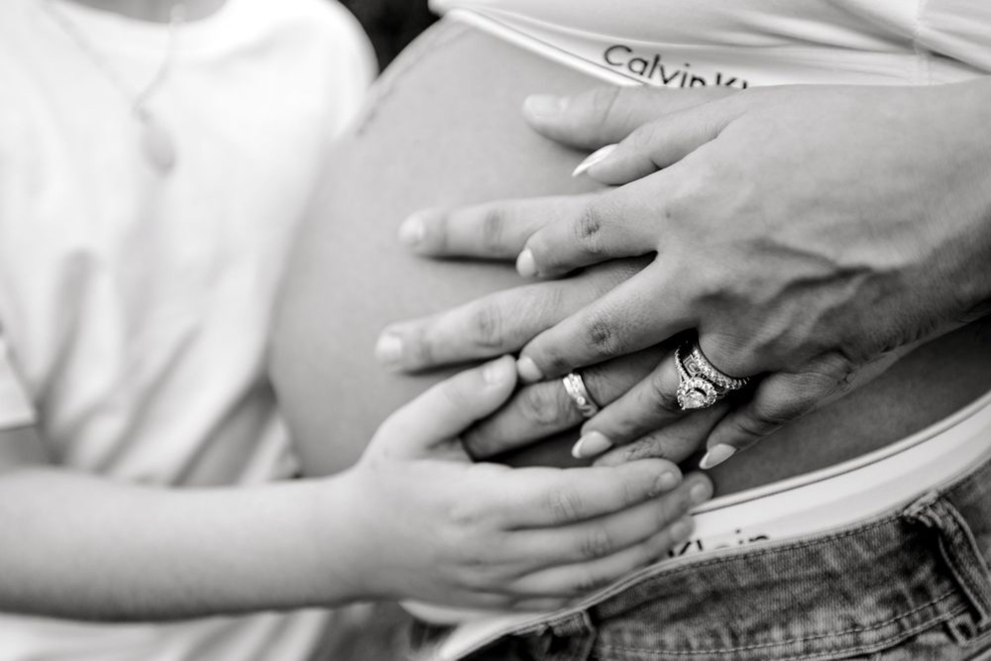 Black-and-white close-up of hands resting on a pregnant belly, wedding rings on display