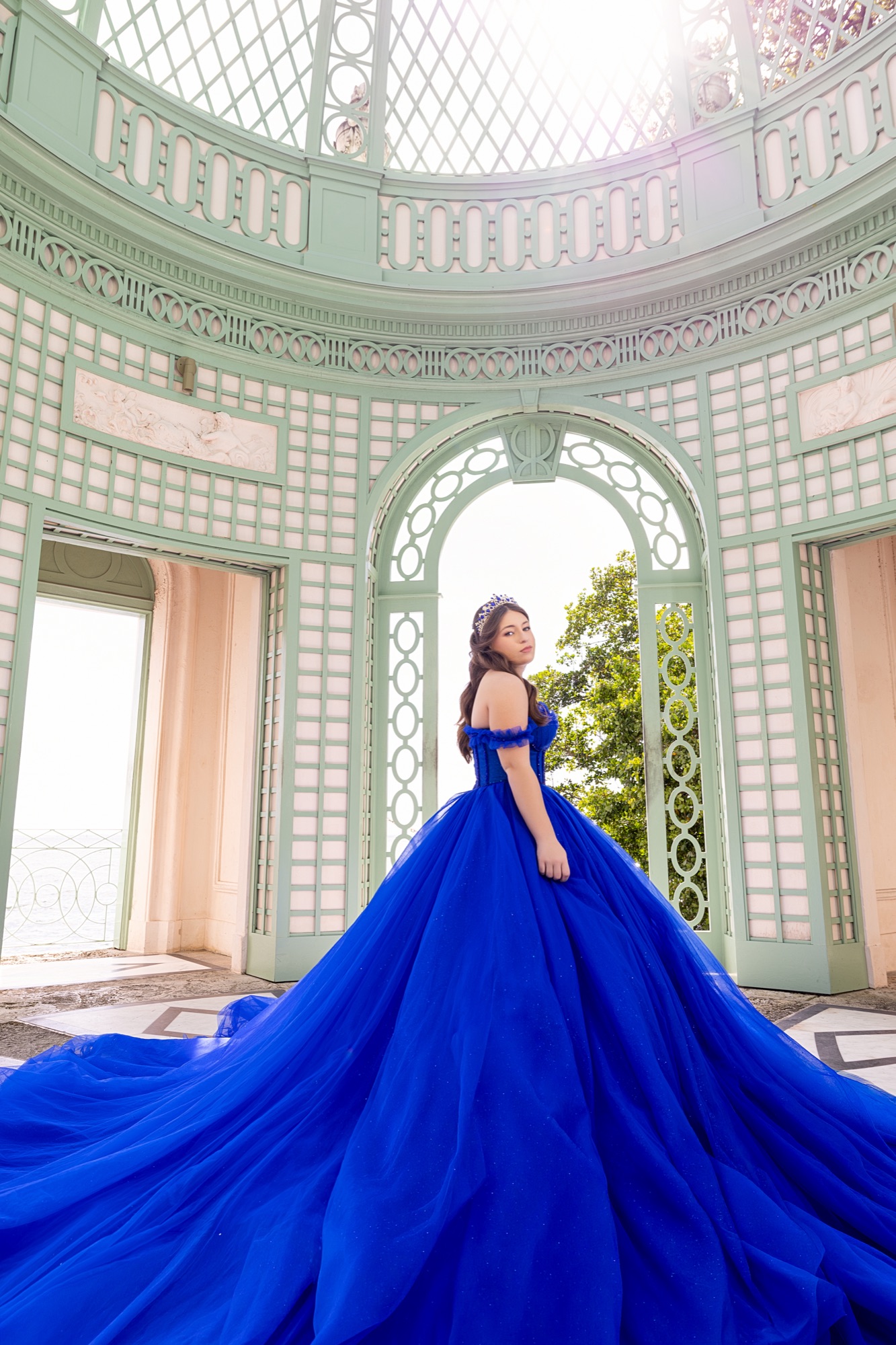 Quinceanera in a royal blue ball gown and crown, her train fanned across a mint lattice pavilion