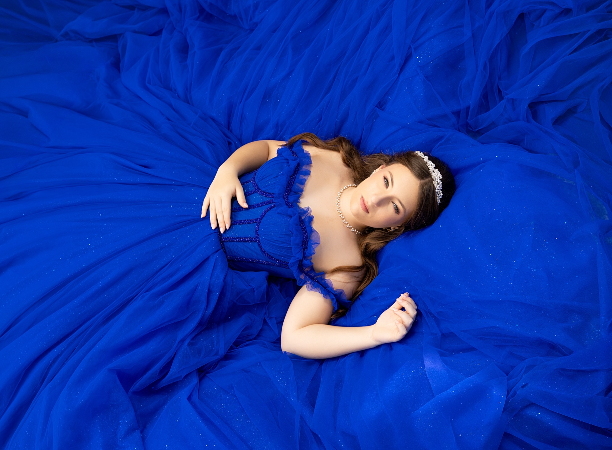 Overhead portrait of a quinceanera lying on her spread royal blue tulle gown