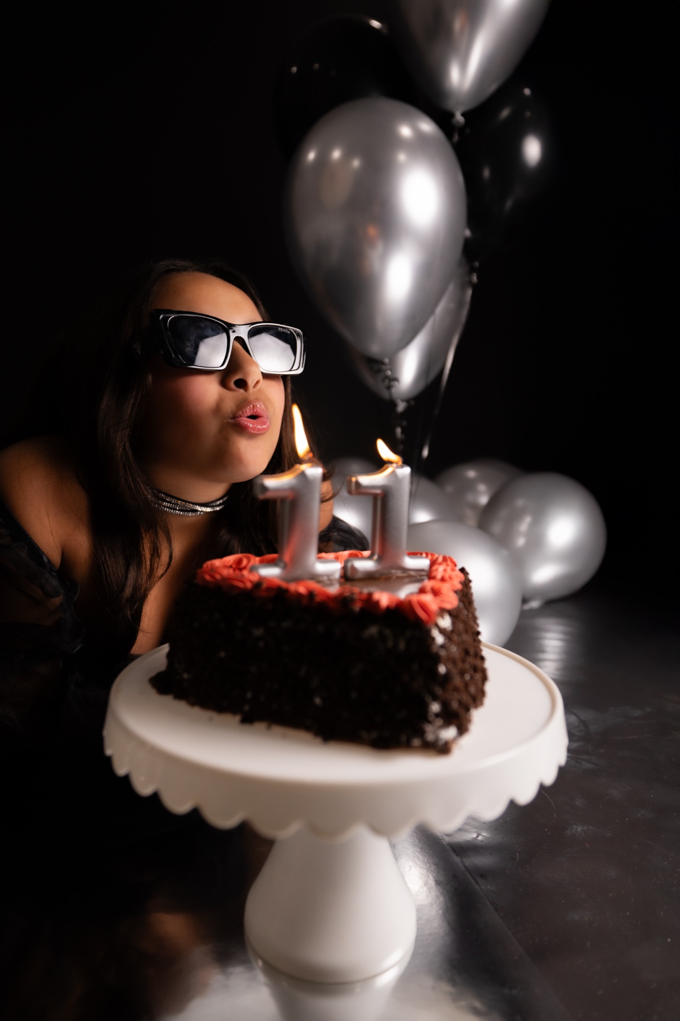 Birthday girl in sunglasses blowing out number 11 candles on a chocolate cake surrounded by silver balloons