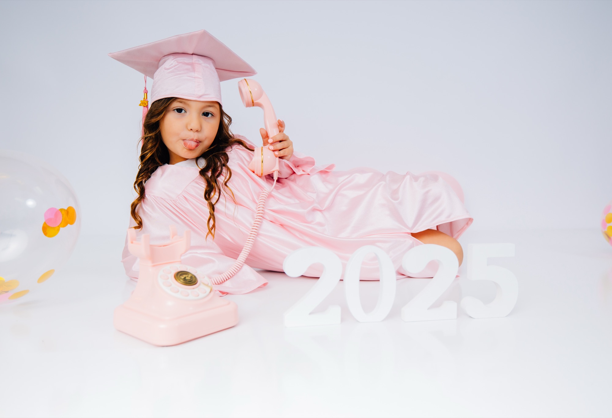 Little graduate in a pink cap and gown posing with a retro phone beside 2025 letters