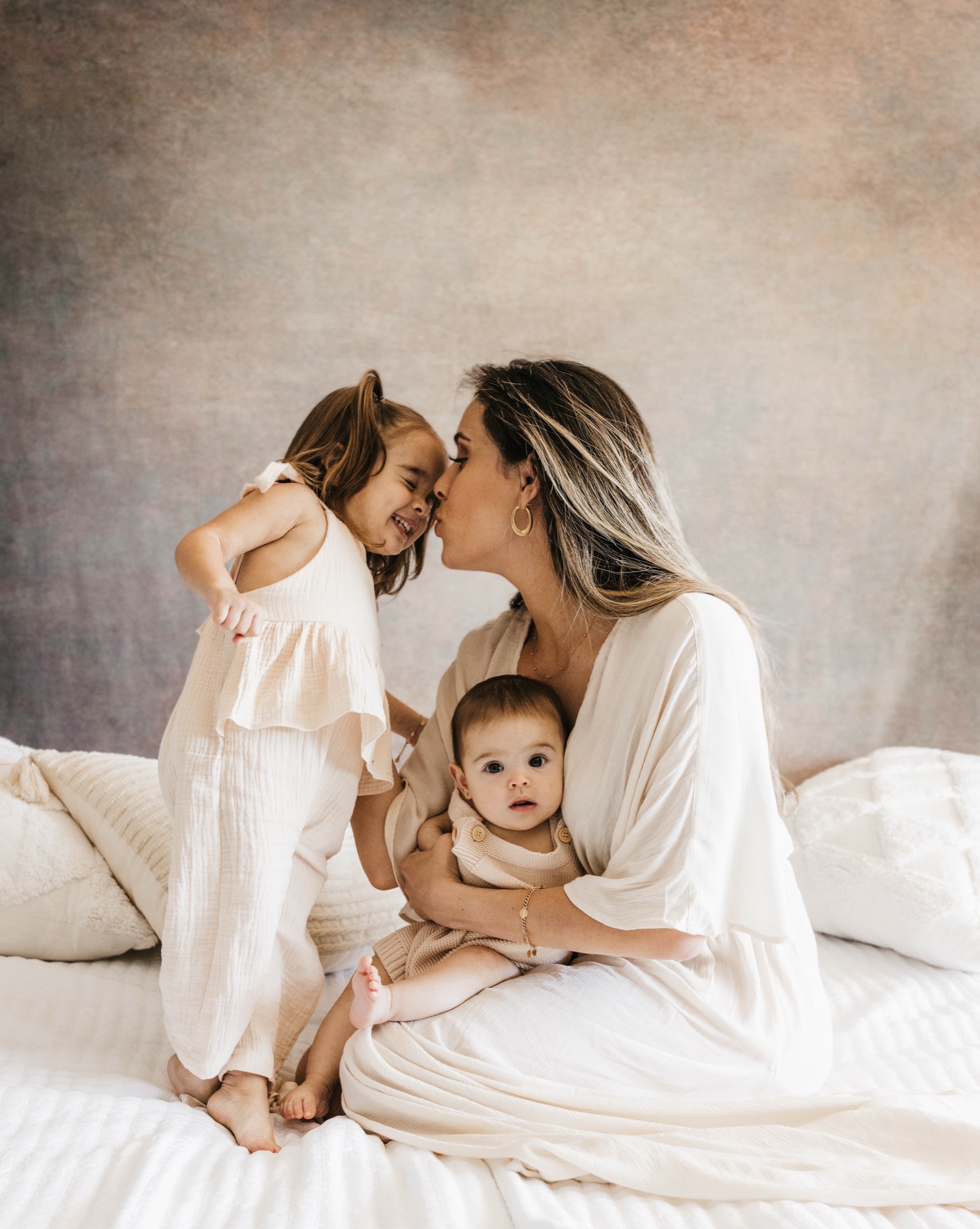 Mother kissing her daughter's forehead while cradling her baby on a soft studio bed set