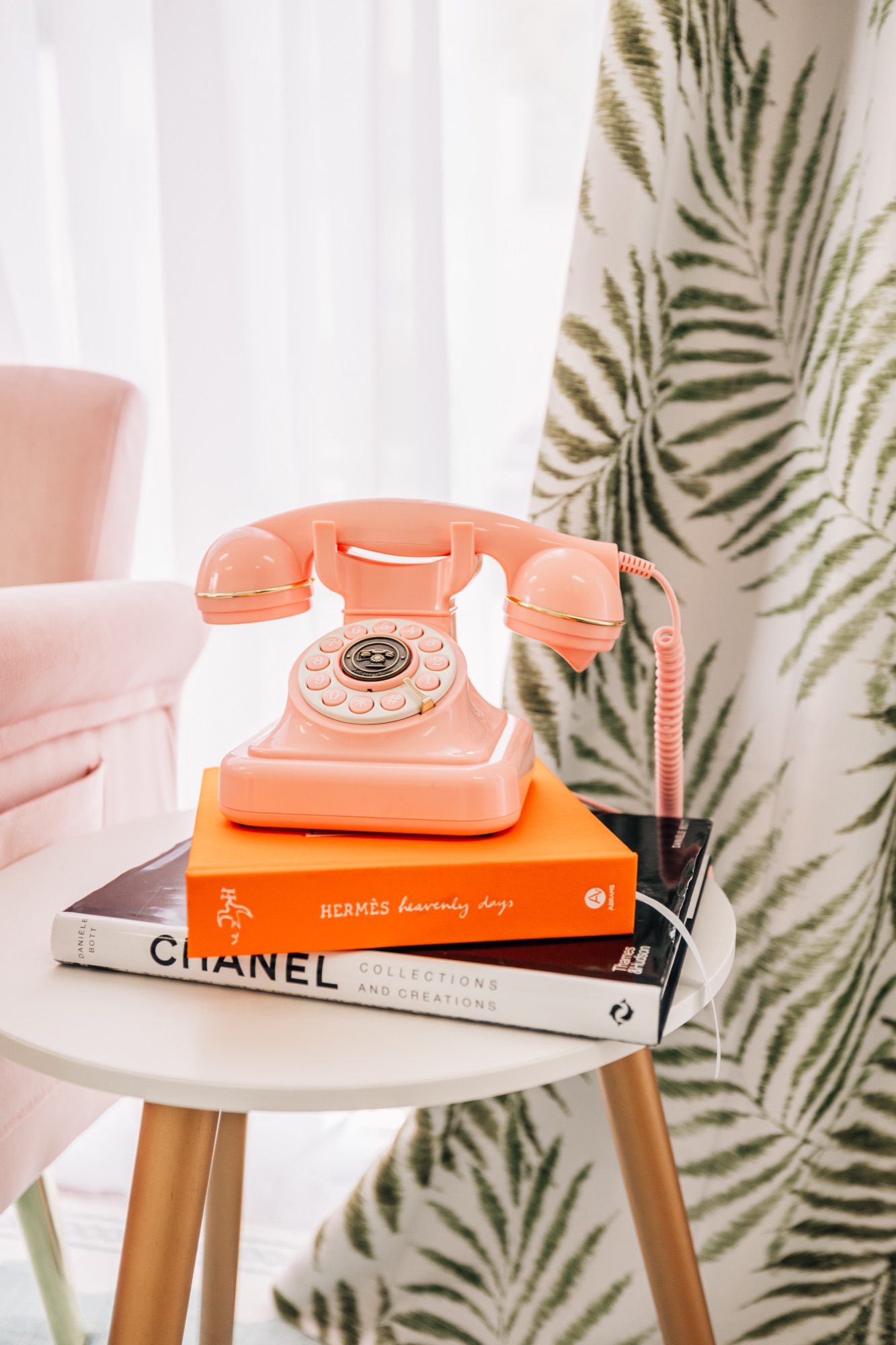Retro pink telephone resting on fashion books beside a palm-print curtain on a styled set